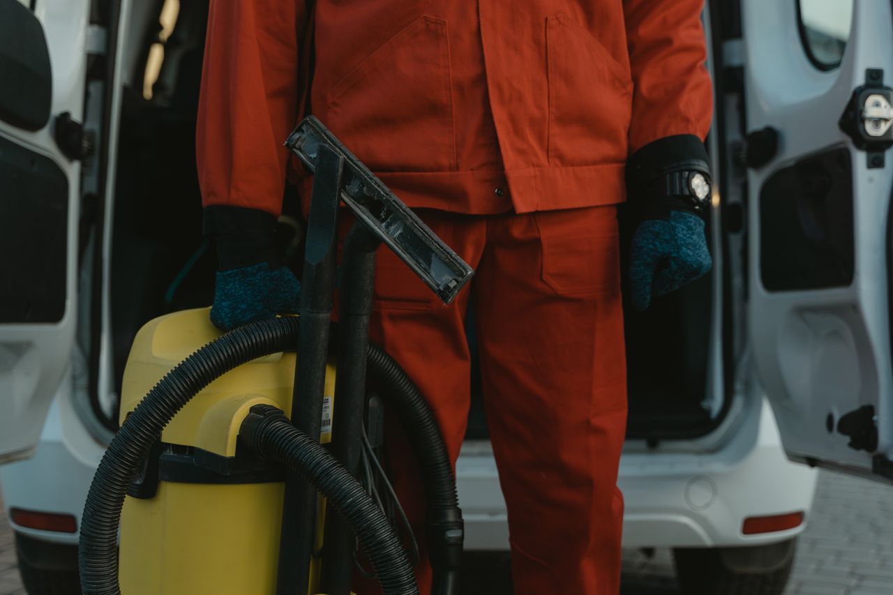 A professional cleaner wearing a red uniform and holding a vacuum beside a van, prepared to provide service.