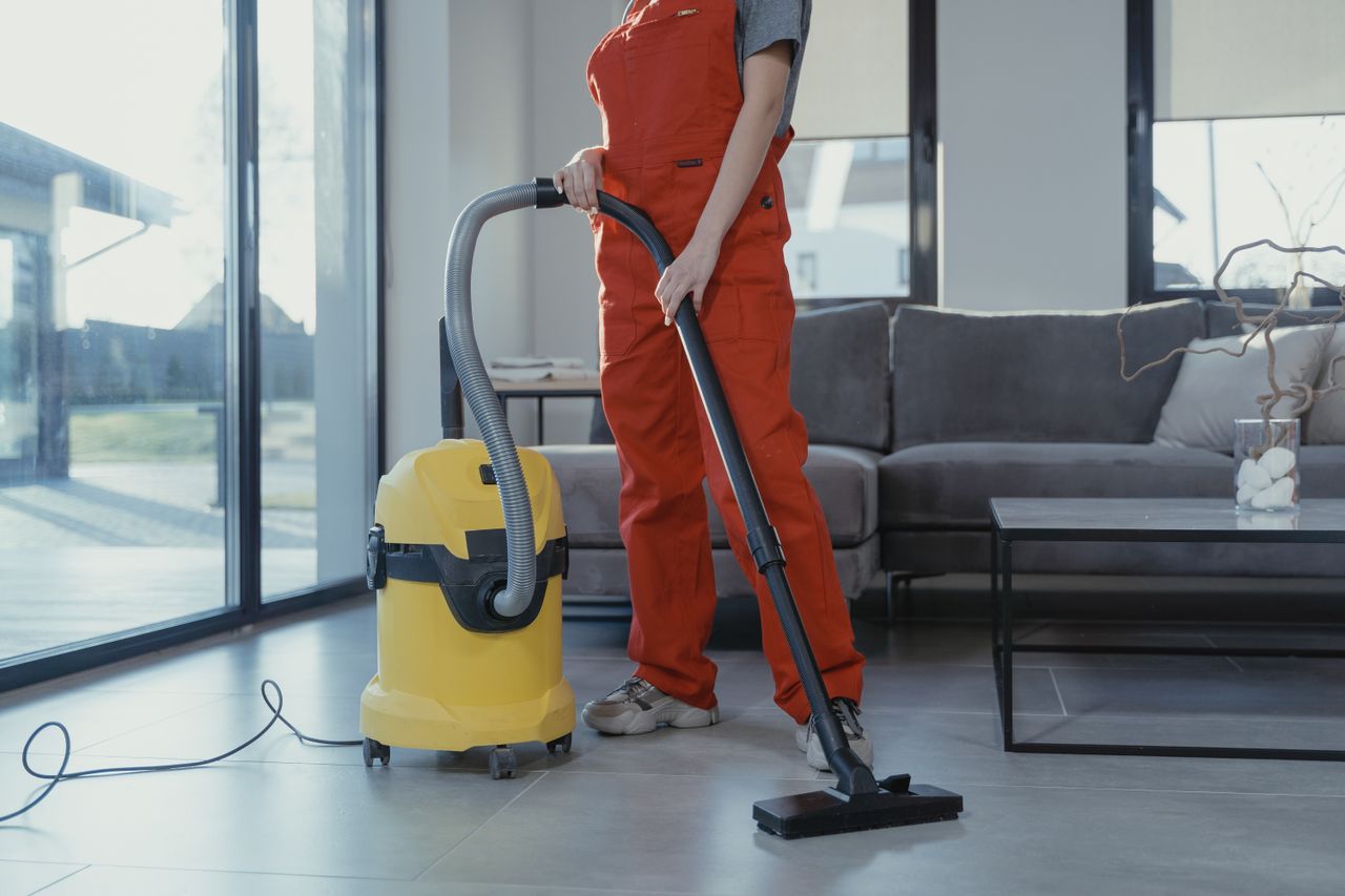 Un professionnel du nettoyage en combinaison rouge aspirant un salon moderne avec un aspirateur jaune.